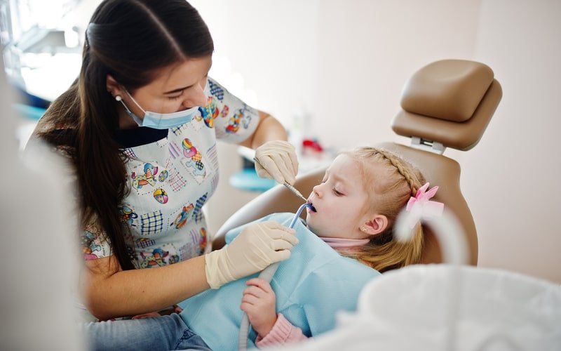 Dentist in a patterned gown examining a young girl's teeth with a mirror